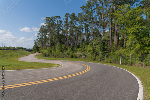 Winding road with double yellow lines in Navarre, Florida. Road near another highway on the left with grass in between and view of a forest trees with metal barriers. © Jason