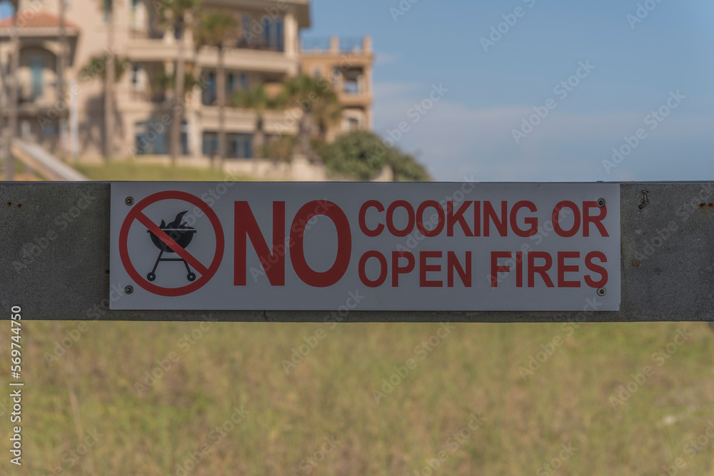 Signage on a wood railing with No Cooking or Open Fire at Destin, Florida outdoors. Close-up of a sign against the grasses and buildings at the blurred background.