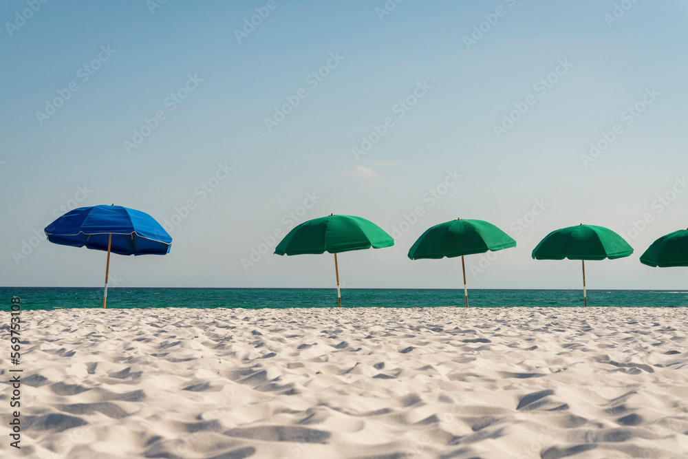 Blue beach umbrella among with the green umbrellas in a row at Destin, Florida. White sand on a beach with beach umbrellas against the ocean below clear sky background.