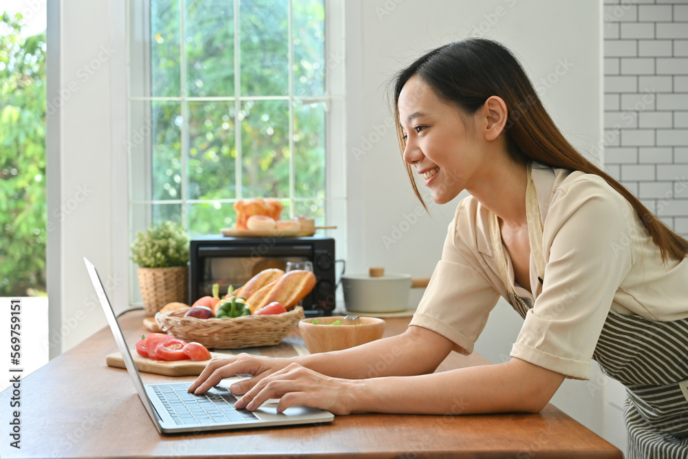 Pretty young asian woman leaning on kitchen table and using laptop, working remote job from home Pretty young asian woman leaning on kitchen table and using laptop, working remote job from home