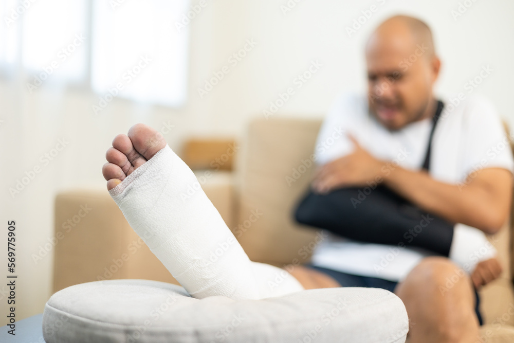 Man suffered pain from accident fracture broken bone injury with leg splints in cast neck splints collar arm splints sling support arm in living room. Social security and health insurance concept. Man suffered pain from accident fracture broken bone injury with leg splints in cast neck splints collar arm splints sling support arm in living room. Social security and health insurance concept.