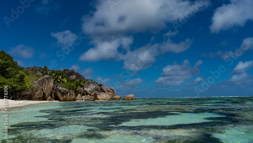 Coral colonies are visible through the clear turquoise water of the ocean. On the sandy beach there are whimsical rounded granite rocks. Tropical vegetation on the hill. Blue sky, clouds. Seychelles