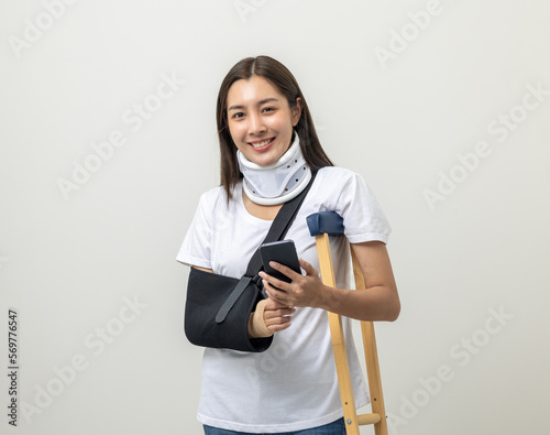 Happy young asian woman broken arm holding smartphone. Woman put on plaster cast splint with walking sticks crutches. Patient wearing sling support arm with neck collar. life insurance and accident
