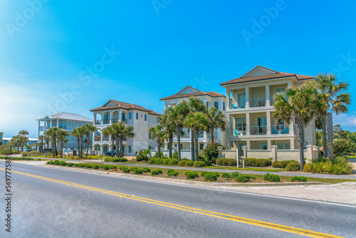Destin, Florida- Fenced beach houses with concrete sidewalks near the highway. There is a highway at the front near the footpath with bushes and palm trees on the side.