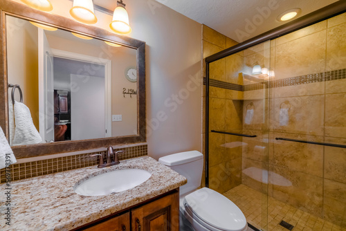 Utah- Small bathroom interior with brown ambience design. There is a vanity sink on the left with towel and framed mirror near the toilet beside shower stall with glass and brown tiles on walls.