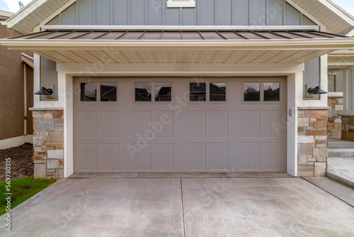Utah- Garage exterior with gray carved canopy garage door with window panes in between wall lights. Attached garage exterior with stone veneer wall and concrete driveway.