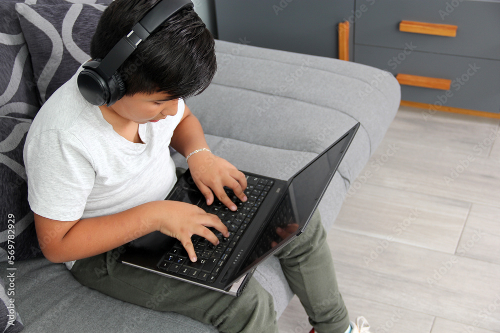 9-year-old Hispanic boy plays video games on his laptop with headphones leading to being overweight and poor sitting posture - obrazy, fototapety, plakaty 