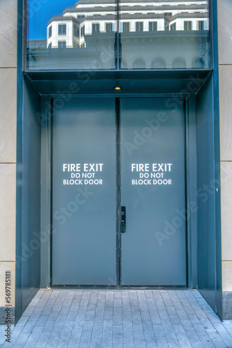 Fototapeta Naklejka Na Ścianę i Meble -  Gray double door with painted Fire Exit Do Not Block Door sign of a building in Austin, Texas. Fire exit door with bricks pathway and glass above with reflection of the building across.