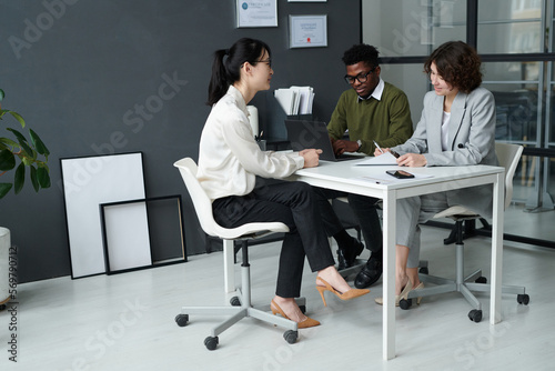 Young woman sitting at table together with managers and talking to them during job interview in modern office
