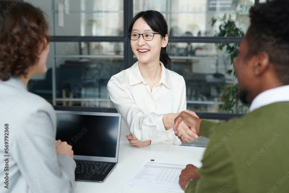Young smiling candidate shaking hands with partners during job interview at table Young smiling candidate shaking hands with partners during job interview at table