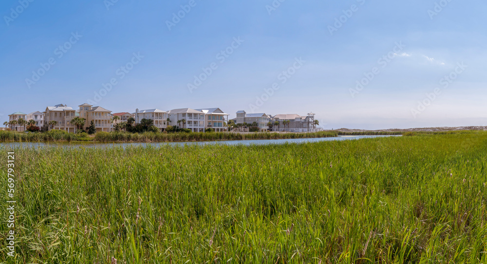 Lake near the three-storey homes on the beach at Destin Point in Destin, Florida. There are tall grasses at the shore of the lake near the houses against the clear skies.