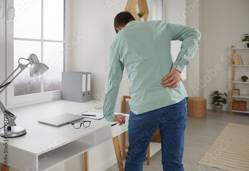 Adult man feels back pain after sedentary work at laptop, sitting at home in uncomfortable position on chair. Tired guy rubs tense back muscles, suffers from pain in the lumbar region of the kidneys.