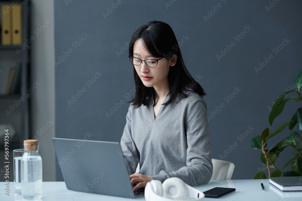 Asian young businesswoman in eyeglasses concentrating on her online work on laptop sitting at her workplace Asian young businesswoman in eyeglasses concentrating on her online work on laptop sitting at her workplace