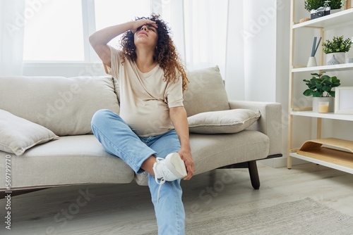 Pregnant woman sitting on the couch at home and wearing pregnancy-friendly shoes with a problem due to the stomach, headache, pregnancy and motherhood difficulties, severe fatigue, headache