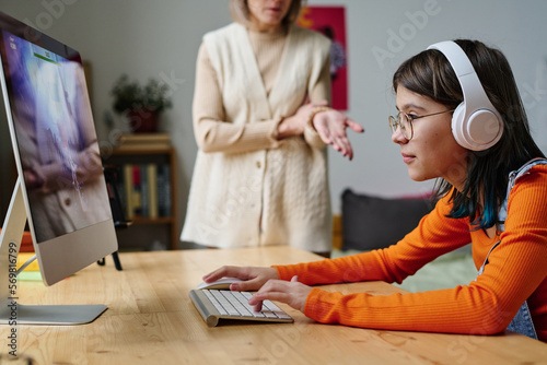 Teenage girl in wireless headphone sitting at table in front of computer monitor and playing computer games with her mother making a remark to her