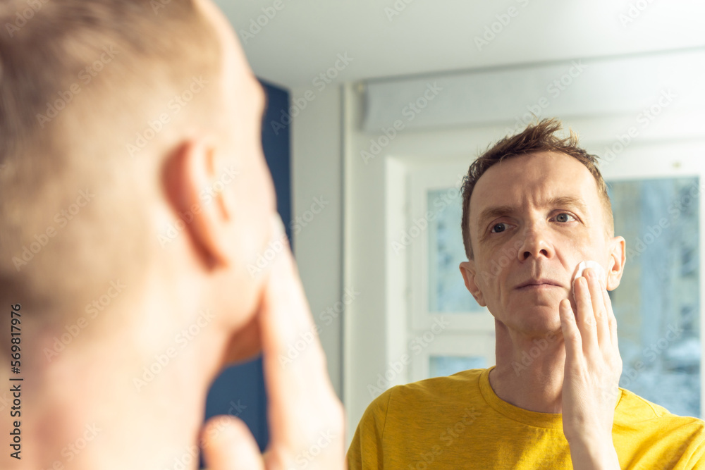 Obraz premium Mature man apply face tonic with cotton pad, then massage with hands. Male portrait in mirror. Skincare morning routine, careful beauty treatment, home lifestyle. Face cleansing, washing.