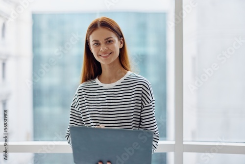 A beautiful young woman with long hair is holding a laptop computer while standing at the window and smiling with her teeth looking at the camera Free Download A beautiful young woman with long hair is holding a laptop computer while standing at the window and smiling with her teeth looking at the camera - GFXLoad