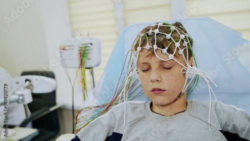 Teenager sits with his eyes closed undergoing EEG testing. Lots of sensors attached to the boy’s head. Close up.