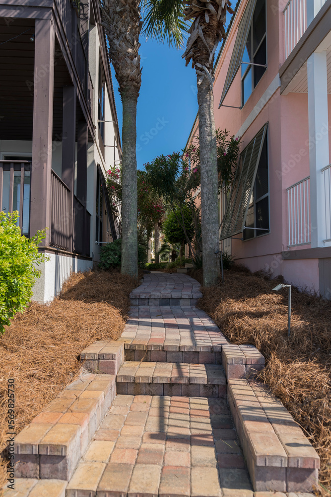 Narrow bricks path with steps in the middle of two houses near Four Prong Lake in Destin, Florida. Walkway with two palm trees and dried grasses at the side of the house buildings in a vertical shot.