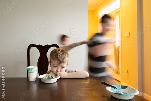 Sad Boy Sits Staring at Table as Brothers Run Around Behind Him