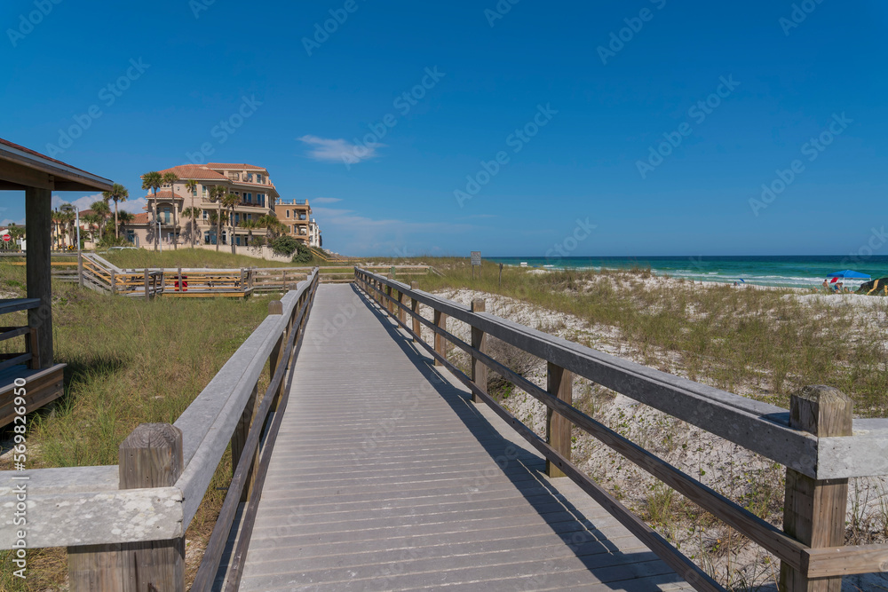 Ocean and beach houses viewed from a wooden pathway in Destin Florida. Scenic coastal landscape with waterfront homes, sandy shore, sea, and blue sky on a sunny day.