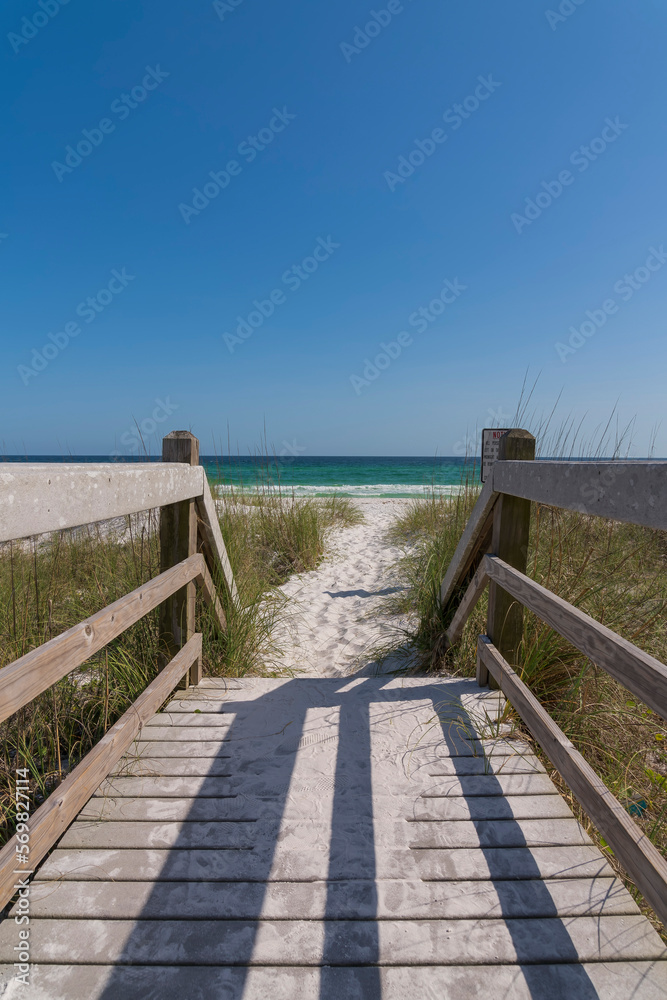 Obraz premium Wooden footbridge with white sand near the stairs heading to the beach at Destin, Florida. Pathway with railings beside the tall grasses on the white sand against the blue ocean and sky background.
