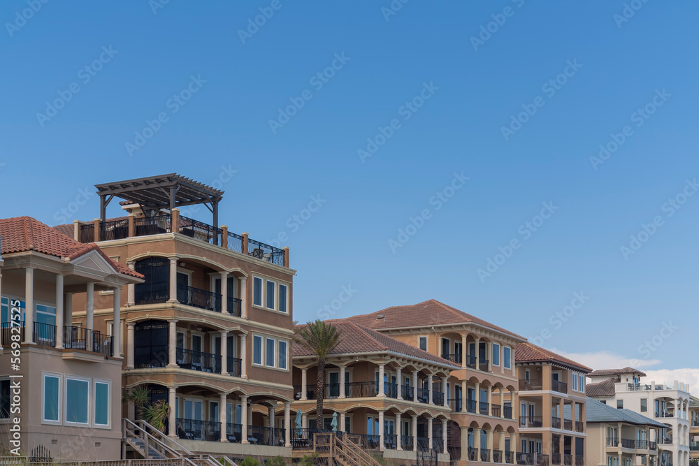 Row of multi-storey beach houses with balconies and roof decks in Destin, Florida. Side view of beach houses' front exterior against the blue sky background. Row of multi-storey beach houses with balconies and roof decks in Destin, Florida. Side view of beach houses' front exterior against the blue sky background.