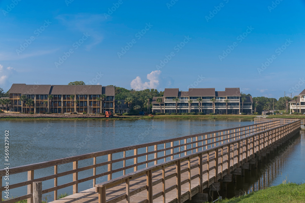 Stewart Lake with footbridge heading to the hotels and apartments on the shore at Destin, Florida. There is a wooden path with railings at the front of low-rise hotel apartments.