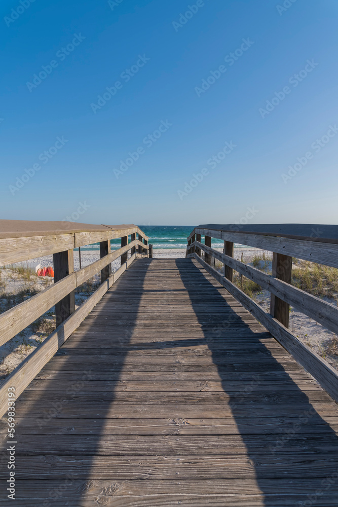 Wooden pathway overlooking ocean and blue sky on a sunny day in Destin Florida. The sunlit walkway leads to the sandy shore and sea with view of the horizon in the distance.
