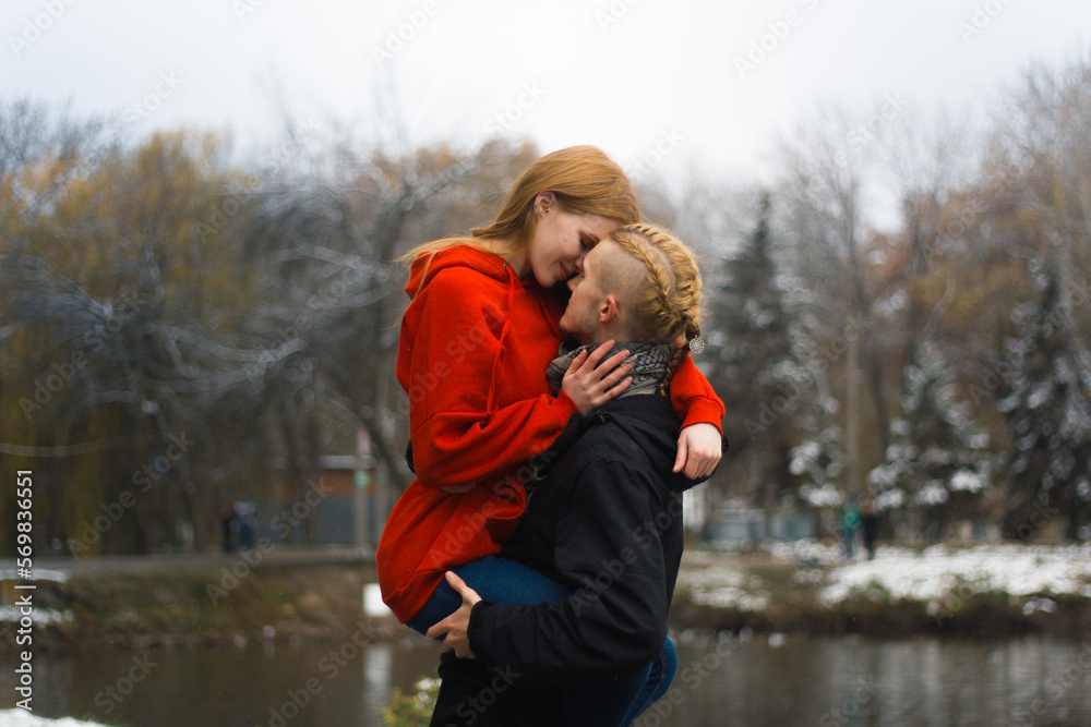 A guy with blond hair holds a red-haired girl in a red sweater in his arms near a winter lake, valentine's day A guy with blond hair holds a red-haired girl in a red sweater in his arms near a winter lake, valentine's day
