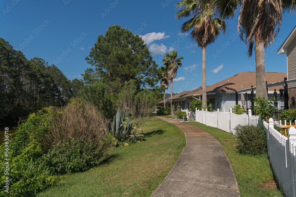 Concrete walkway on a grass with bushes outside the fenced residences in Navarre, Florida. There are plants and forest with tall trees on the left near the houses with white picket fence on the right.