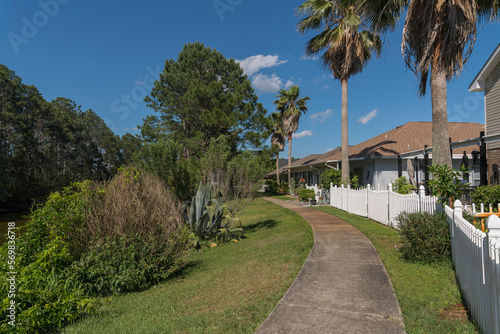 Concrete walkway on a grass with bushes outside the fenced residences in Navarre, Florida. There are plants and forest with tall trees on the left near the houses with white picket fence on the right.