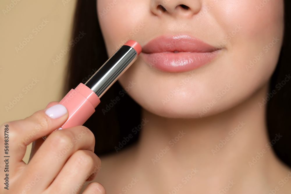 Young woman applying nude lipstick on beige background, closeup