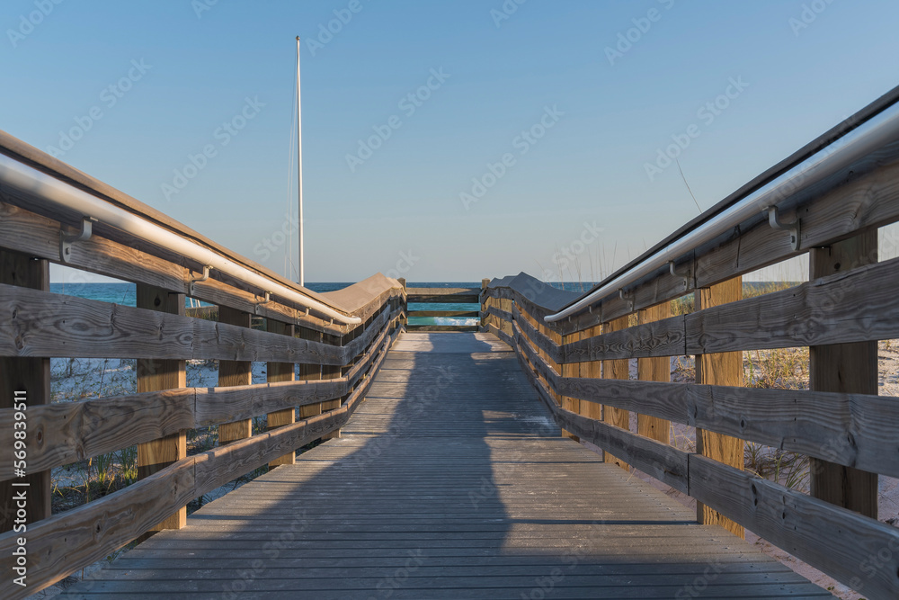 Wooden pathway with metal handrails mounted on railings at the beach in Destin, Florida. Pathway heading to the beach with views of a flag pole on the left and clear skyline background.