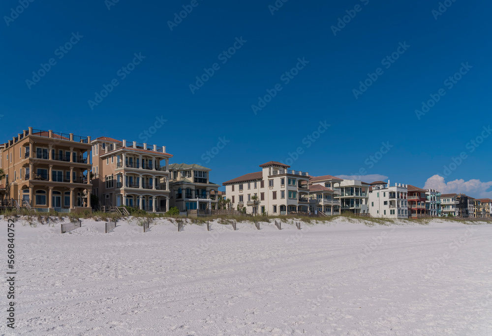 Beach houses with roof decks and balconies facing the shore of the beach in Destin, Florida. There are fences of sand dunes with grasses with wooden footbridges at front of houses against the sky.