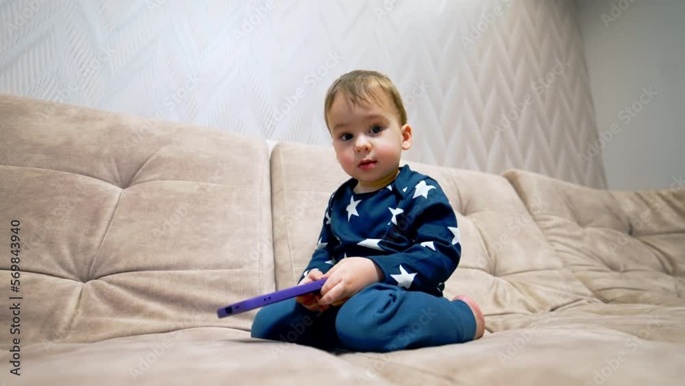 Adorable one year old toddler playing with phone sitting on sofa. Kid drops the gadget and touches camera. Low angle view close up. Adorable one year old toddler playing with phone sitting on sofa. Kid drops the gadget and touches camera. Low angle view close up.