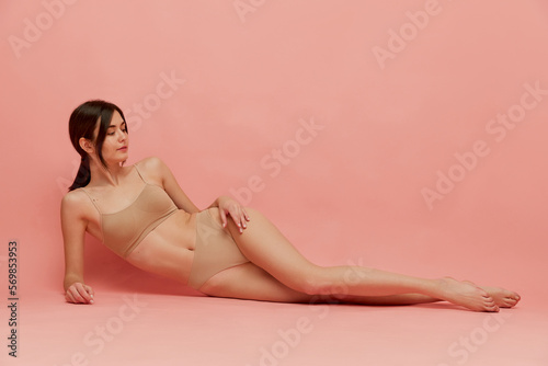 Spa. Beautiful young woman with slim body shape lying on floor, posing in beige underwear posing over pink studio background. Concept of body and skin care, fitness, natural beauty, health, wellness.