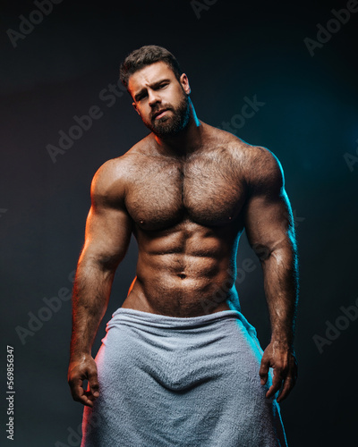 Big muscled man in white towel. Handsome hunk posing is studio. Naked male model with muscles and hairy body at dark background. Bodybuilder wearing only bath towel. Sexy athlete with six pack abs.