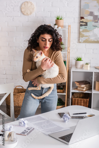 Copywriter holding oriental cat near devices and documents on table.