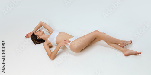 Portrait of young beautiful girl with slim body posing in white cotton underwear posing on floor over grey studio background. Concept of body and skin care, fitness, natural beauty, health, wellness.