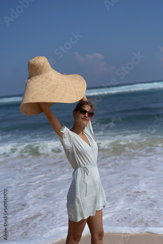 Young happy smile woman on the ocean. A beautiful woman in a straw hat and a sundress walks along the ocean shore barefoot on the sand.