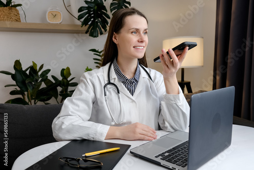 Leinwand Poster Smiling beautiful young woman doctor recording audio message on smartphone sitti