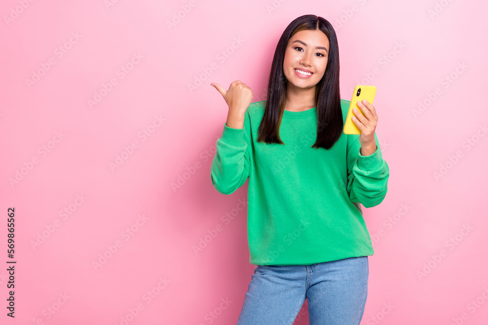Photo of young cheerful positive promoter korean lady wear stylish outfit hold phone directing finger empty space isolated on pink color background Photo of young cheerful positive promoter korean lady wear stylish outfit hold phone directing finger empty space isolated on pink color background