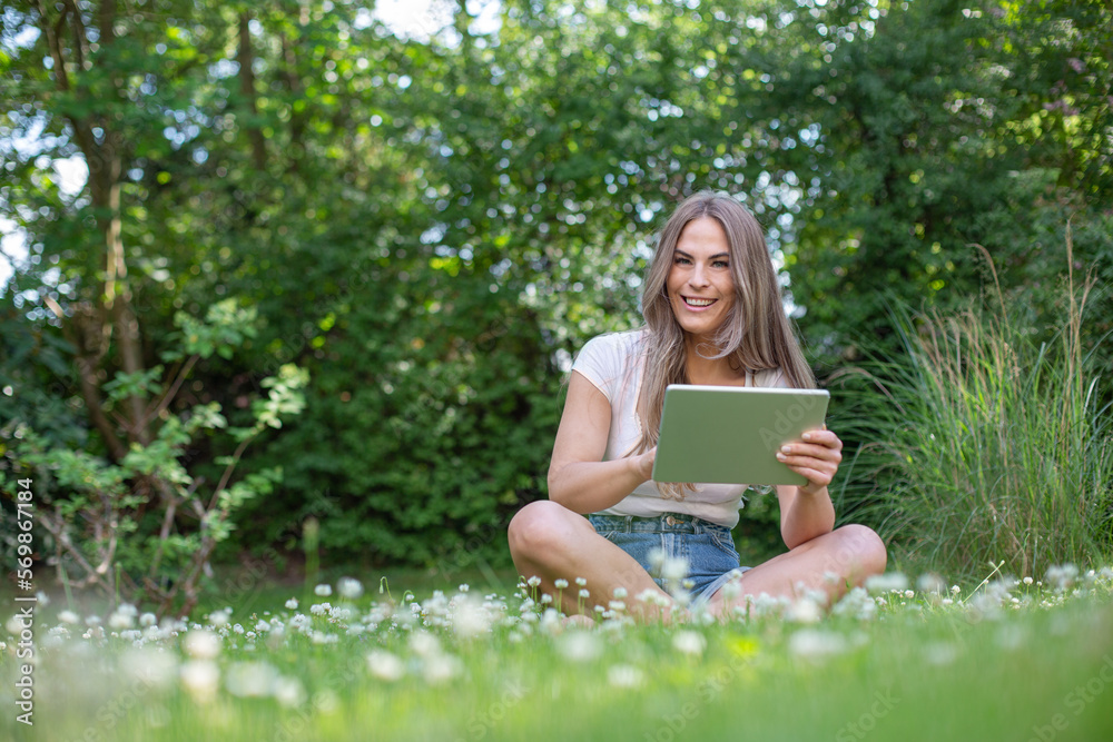 Young pretty blonde woman sitting in the garden with an tablet and reading, playing, online banking or using an app Young pretty blonde woman sitting in the garden with an tablet and reading, playing, online banking or using an app