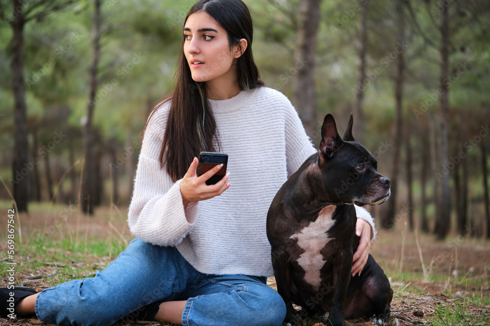 Young caucasian woman using the smartphone with her american staffordshire and french bulldog mixed breed dog sitting at a pine forest. Young caucasian woman using the smartphone with her american staffordshire and french bulldog mixed breed dog sitting at a pine forest.