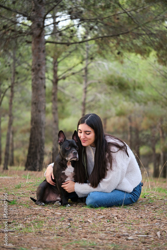 Young caucasian woman hugging her american staffordshire and french bulldog mixed breed dog at a pine forest.