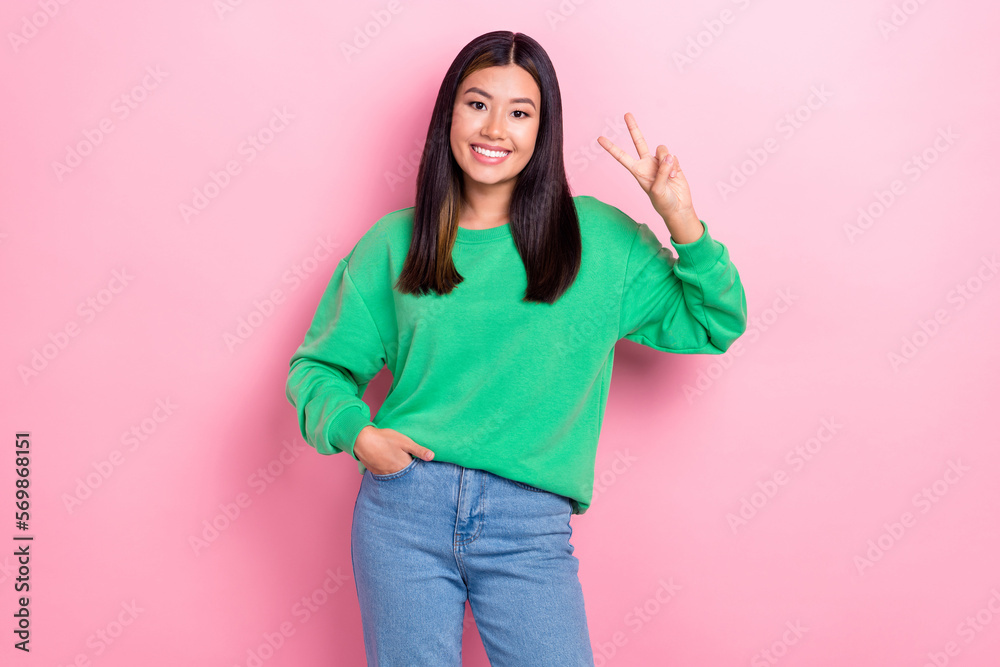 Photo of cheerful positive chinese lady wear green pullover with denim jeans showing v-sign hello peace everyone isolated on pink color background Photo of cheerful positive chinese lady wear green pullover with denim jeans showing v-sign hello peace everyone isolated on pink color background