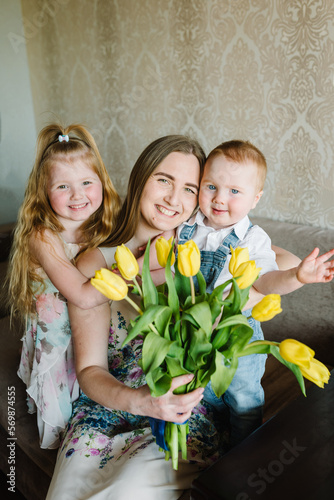 Mom, son and daughter smiling and hugging. Mother's day concept. Girl and boy congratulates mother and gives a bouquet of flowers tulips at home. Holiday greeting card for International Women's Day.