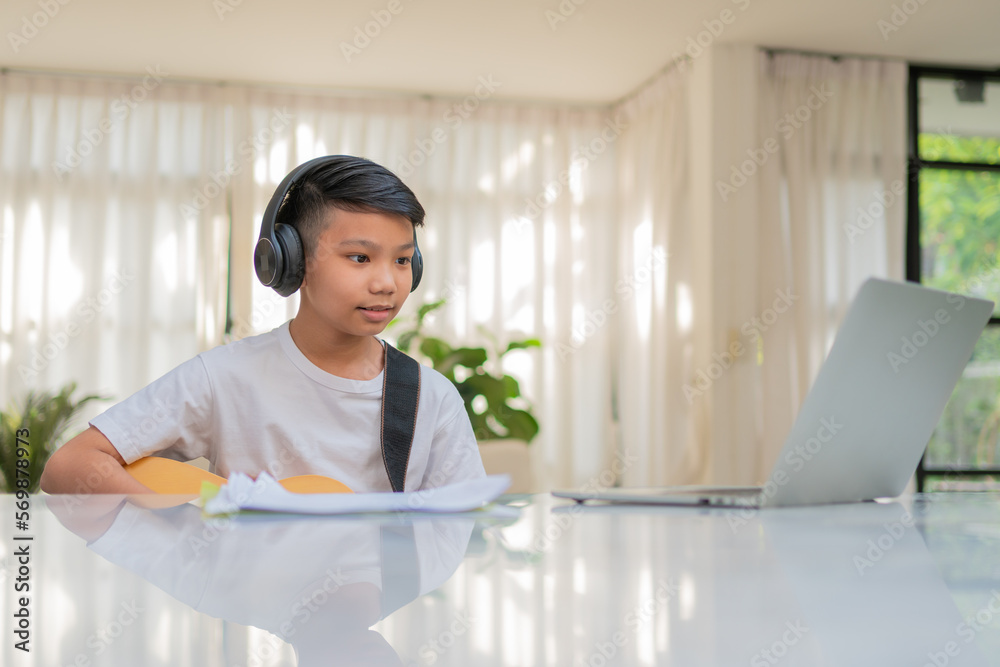 Asian boy playing guitar and watching online course on laptop while practicing for learning music or musical instrument online at home. Boy students study online with video call teachers play guitar.