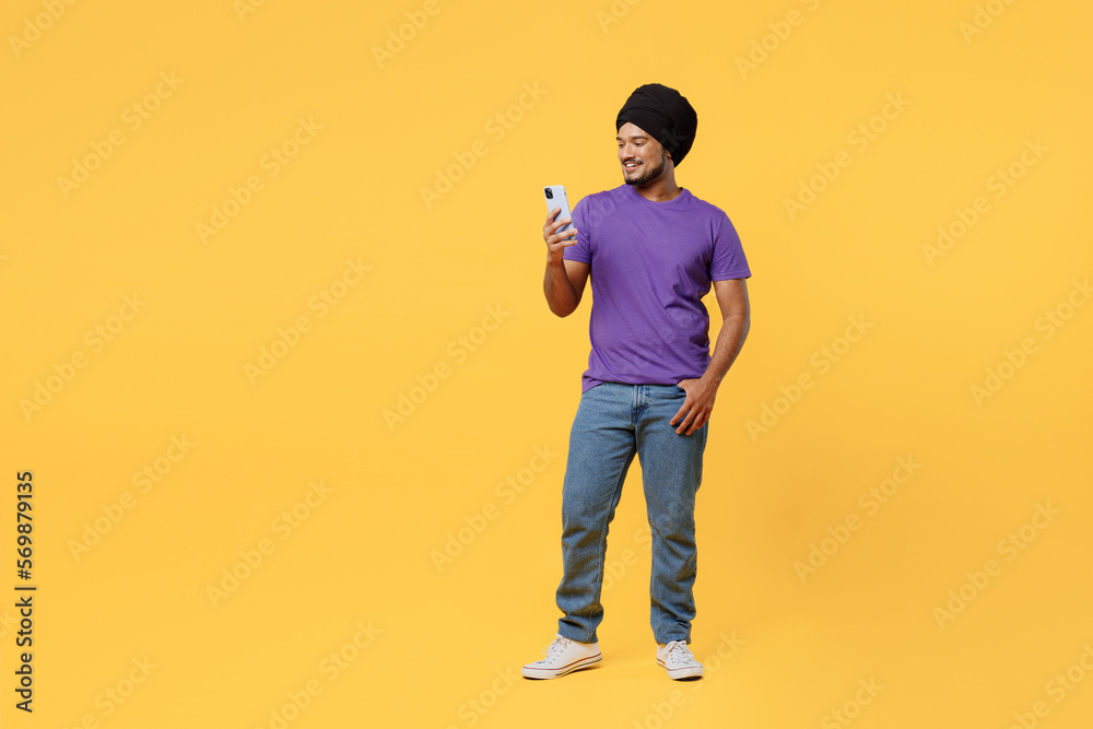 Full body happy devotee Sikh Indian man ties his traditional turban dastar wear purple t-shirt hold in hand use mobile cell phone chatting online isolated on plain yellow background studio portrait. Full body happy devotee Sikh Indian man ties his traditional turban dastar wear purple t-shirt hold in hand use mobile cell phone chatting online isolated on plain yellow background studio portrait.